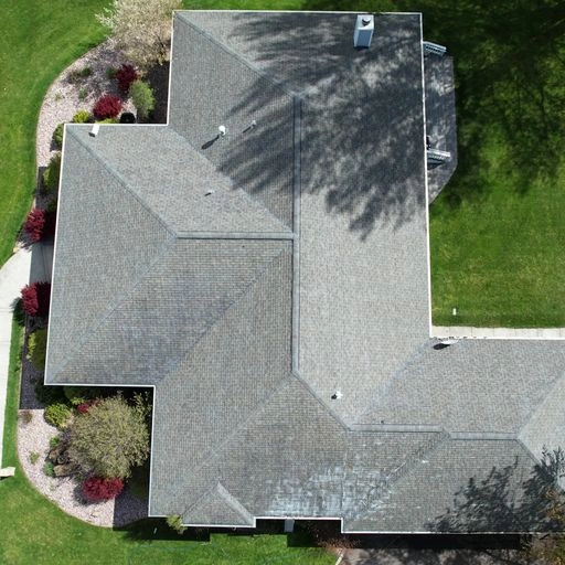 Aerial view of a house with a gray shingle roof, surrounded by green grass, landscaping with bushes and trees, and a concrete pathway on the right.