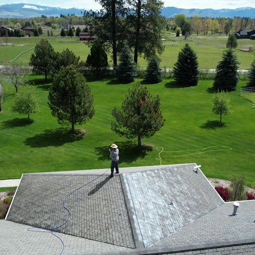 A person stands on a roof, power washing shingles. The roof is partially cleaned, with visible color contrast. Green trees, a grassy yard, and distant mountains are in the background under a partly cloudy sky.