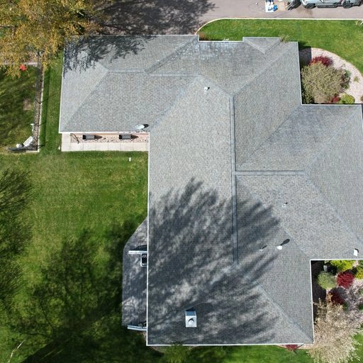 Aerial view of a house with a gray shingled roof, surrounded by green lawn, trees casting shadows, and landscaped garden areas along the edges.