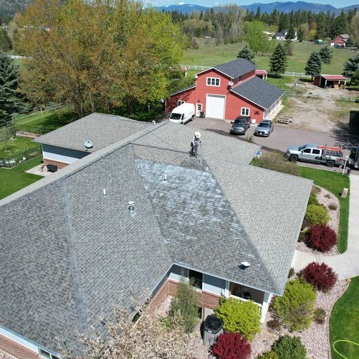 A person is working on the roof of a house, which is surrounded by a well-kept yard with trees and bushes. In the background, there are red barns, vehicles, and mountains under a partly cloudy sky.