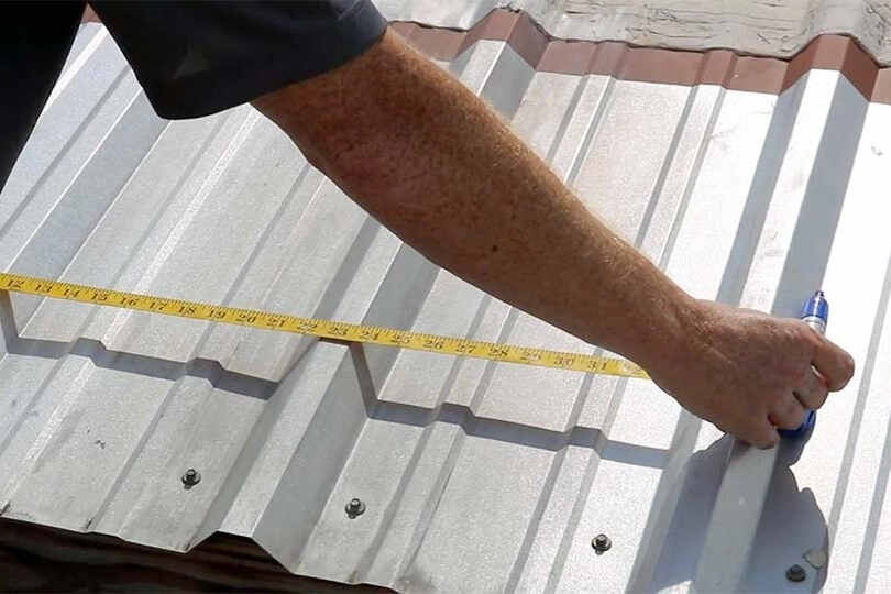 A person measures a corrugated metal roof panel with a yellow measuring tape, holding a blue marker and marking a measurement on the panel.