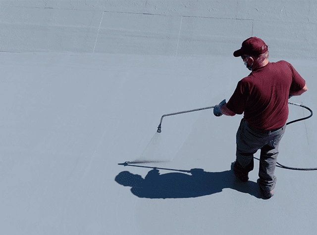 A person in a maroon shirt, cap, and gloves sprays a gray coating onto a flat surface with a long-handled sprayer, casting a shadow on the coated area.