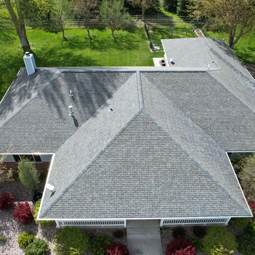 Aerial view of a house with a gray shingled roof, surrounded by green lawn, trees, and landscaped bushes along a walkway leading to the entrance.
