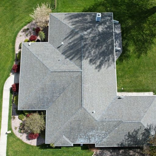 Aerial view of a large, gray-roofed house surrounded by well-manicured green lawns, landscaped bushes, and trees casting shadows on the roof and yard.