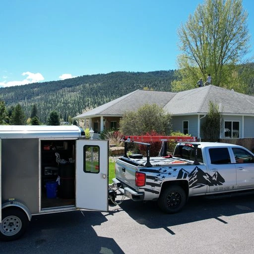 A white pickup truck with a mountain graphic is parked beside a house with mountains in the background. The truck is towing an open trailer filled with equipment. The sky is mostly clear with a few clouds.